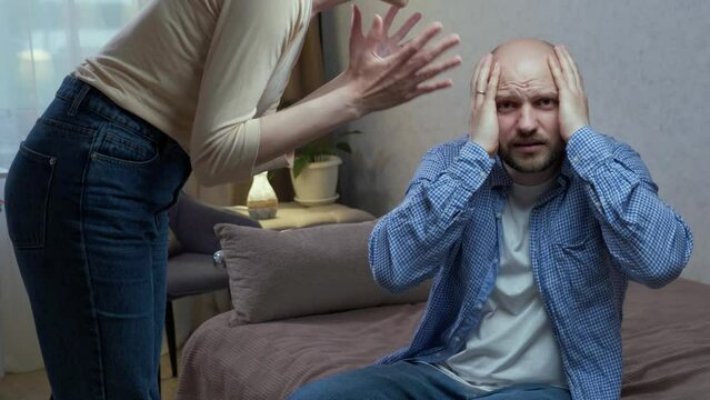 Abuse in the family and psychological pressure. The evil tyrant wife stands and shouts at her husband. The frightened husband holds his head with his hands and looks at the camera