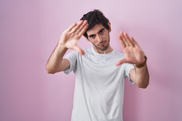 Young hispanic man standing over pink background doing frame using hands palms and fingers, camera perspective