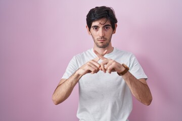 Young hispanic man standing over pink background rejection expression crossing fingers doing...