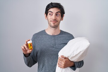 Young hispanic man wearing pyjama hugging pillow holding pills smiling looking to the side and staring away thinking.