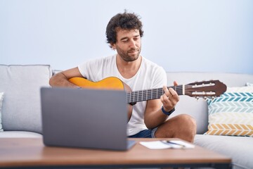 Young hispanic man having online classical guitar class sitting on sofa at home