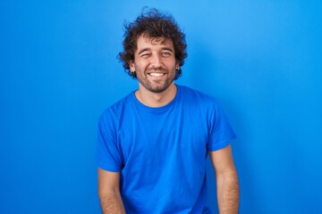 Hispanic young man standing over blue background winking looking at the camera with sexy expression, cheerful and happy face.