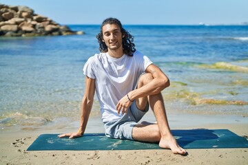 Young hispanic man doing yoga exercise sitting on sand at beach