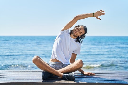 Young hispanic man doing yoga exercise sitting on bench at seaside