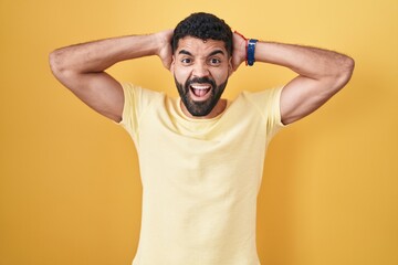 Hispanic man with beard standing over yellow background crazy and scared with hands on head, afraid and surprised of shock with open mouth