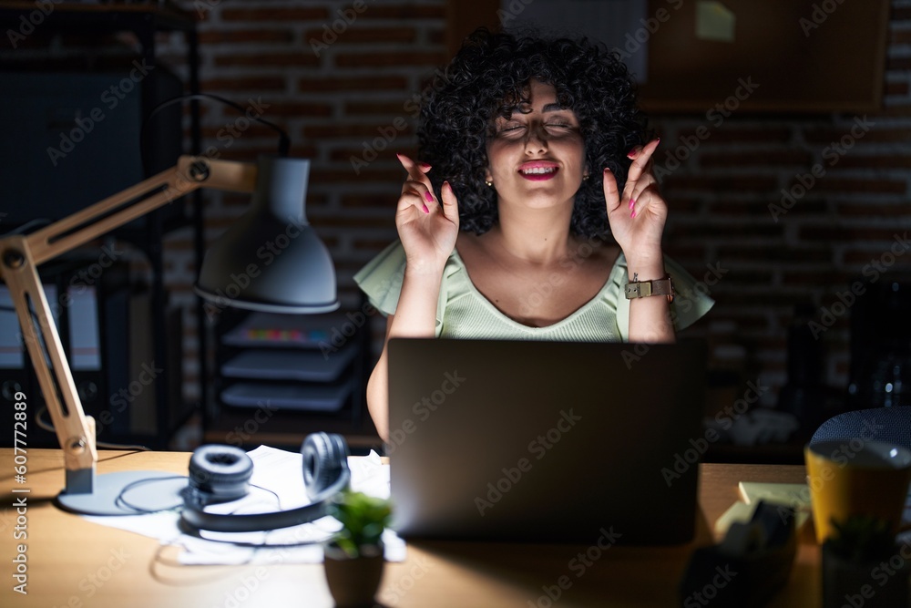 Poster Young brunette woman with curly hair working at the office at night gesturing finger crossed smiling with hope and eyes closed. luck and superstitious concept.