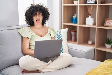 Fototapeta na wymiar Young brunette woman with curly hair using laptop sitting on the sofa at home sticking tongue out happy with funny expression. emotion concept.