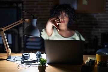 Young brunette woman with curly hair working at the office at night looking unhappy and angry showing rejection and negative with thumbs down gesture. bad expression.