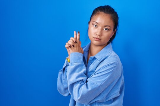 Asian young woman standing over blue background holding symbolic gun with hand gesture, playing killing shooting weapons, angry face