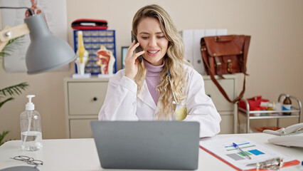 Young blonde woman doctor using laptop talking on smartphone at clinic