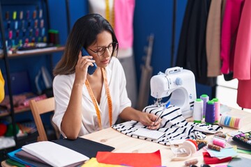Fototapeta premium Young hispanic woman tailor talking on smartphone writing on notebook at sewing studio
