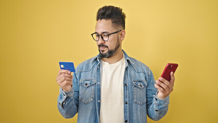 Young latin man shopping with smartphone and credit card over isolated yellow background