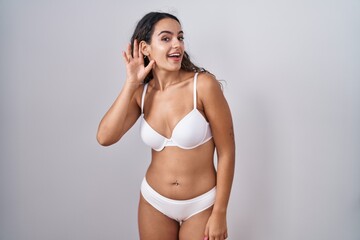 Young hispanic woman wearing white lingerie smiling with hand over ear listening an hearing to rumor or gossip. deafness concept.