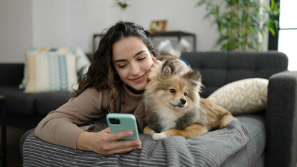 Young hispanic woman with dog using smartphone lying on sofa at home