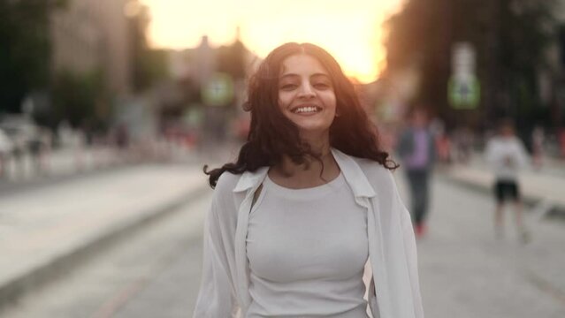 Charming young indian woman walking down the street turns around with flying hair and looking at camera outdoors Happy relaxed lady walking on the city centre enjoying beautiful sunset alone