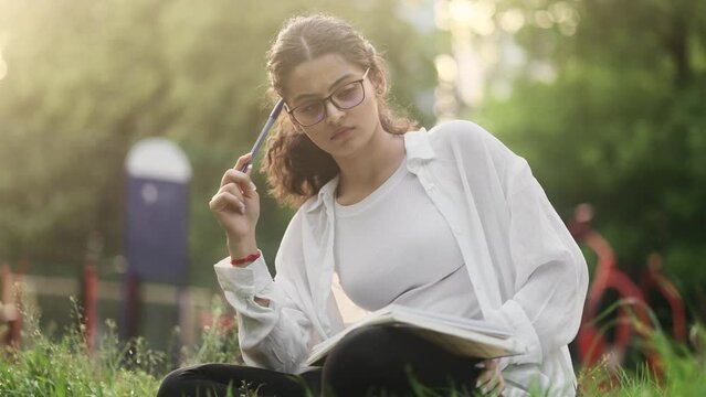 Pretty indian girl student thinks questioningly and finding solution idea while write homework make notes sitting on grass at park Charming young curly woman has remote distance education outdoors
