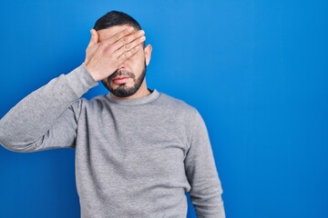 Hispanic man standing over blue background covering eyes with hand, looking serious and sad....