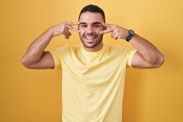 Fototapeta na wymiar Young hispanic man standing over yellow background doing peace symbol with fingers over face, smiling cheerful showing victory