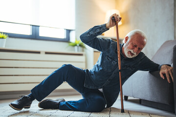 Elder senior man lying on floor after falling down with wooden walking stick beside couch on rug in...