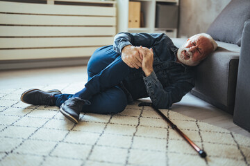 Elder senior man lying on floor after falling down with wooden walking stick beside couch on rug in...