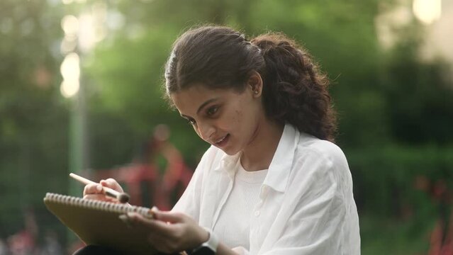 Portrait of pretty young indian woman illustrator drawing sketch from nature using brush or pencil outdoors Pretty curly female enjoying beautiful sunny day while painting in green park Art hobby