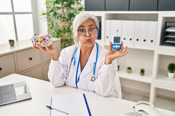 Middle age woman with grey hair wearing doctor uniform holding glucose monitor puffing cheeks with funny face. mouth inflated with air, catching air. - obrazy, fototapety, plakaty