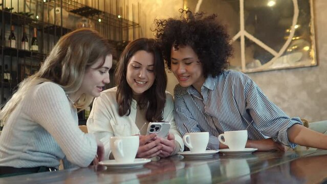 Three multiethnic young women friends in a coffee shop laughing and talking while watching the smart phone