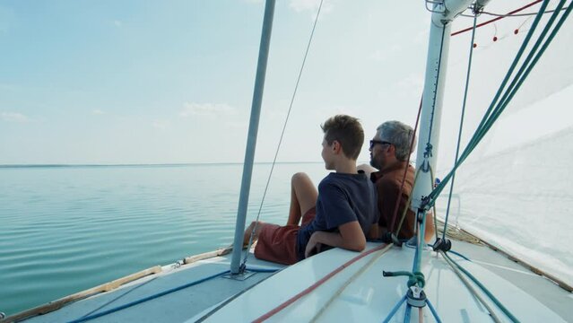Modern mature man and his teen son sitting relaxed on board the sailboat talking about their travel