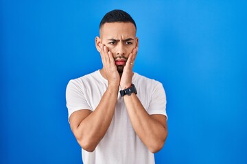 Young hispanic man standing over blue background tired hands covering face, depression and sadness, upset and irritated for problem