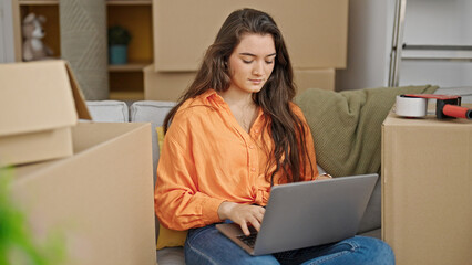 Young beautiful hispanic woman using laptop sitting on sofa at new home