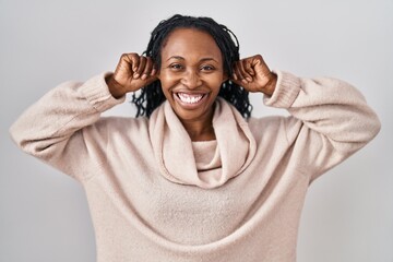 African woman standing over white background smiling pulling ears with fingers, funny gesture. audition problem