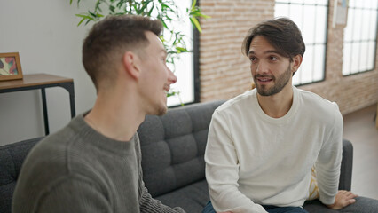 Two men couple sitting on sofa speaking at home