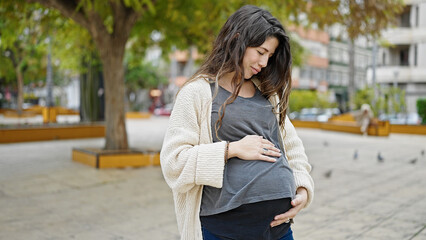 Young pregnant woman standing with serious expression touching belly at park