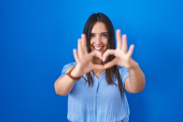 Young brunette woman standing over blue background smiling in love doing heart symbol shape with hands. romantic concept.