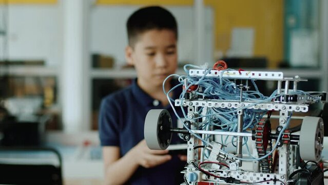Schoolboy assembling the complicated moving robot in a class at a robotics lesson and trying how the wheels moves with a joystick. Concept of technological modern education