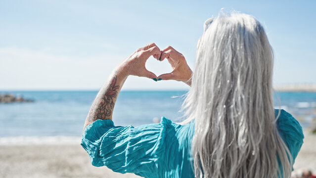 Middle age grey-haired woman doing heart gesture with hands at beach