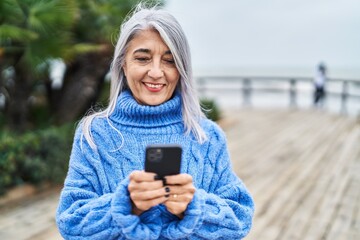 Middle age grey-haired woman smiling confident using smartphone at seaside