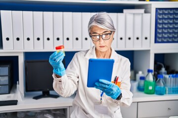 Middle age grey-haired woman scientist using touchpad holding test tube at laboratory