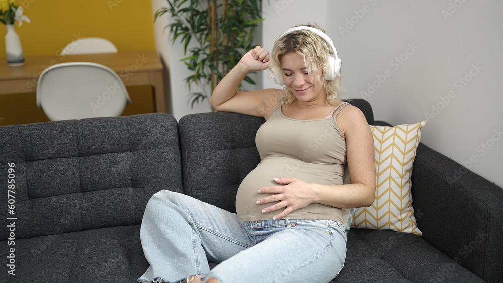 Poster Young pregnant woman listening to music touching belly and dancing at home