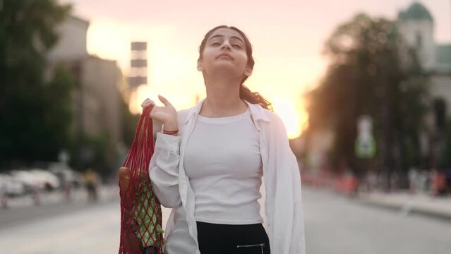 Charming young indian woman with eco bag walking down the street and looking around at evening after supermarket outdoors Happy relaxed lady walking on the city centre enjoying beautiful sunset alone