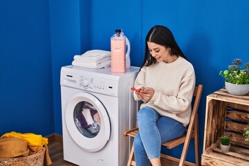 Young beautiful hispanic woman using smartphone waiting for washing machine at laundry room