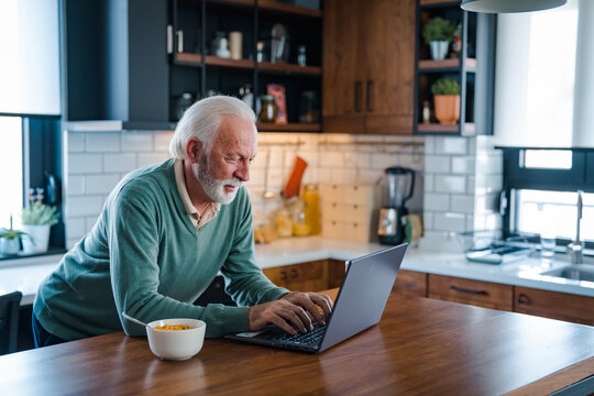 Senior man working at laptop at home. Retired Man On Phone At Home In Kitchen Using Laptop Celebrating Good News. Senior adult in home kitchen with tea cup and laptop.