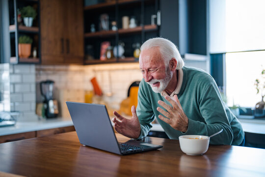 Cheerful senior man during video conference in kitchen on the laptop while enjoying breakfast and a cup of coffee. Elderly person using internet online chat technology video webcam making a video call