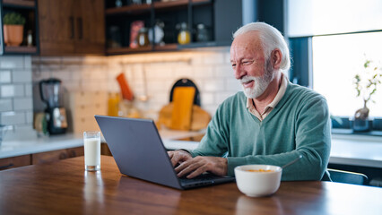 Cheerful senior man during video conference in kitchen on the laptop while enjoying breakfast and a...