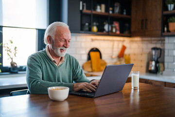 Fototapeta premium One senior businessman standing in the kitchen and working on the laptop while drinking coffee. Grey-haired bearded senior man eyeglasses looking concentrated