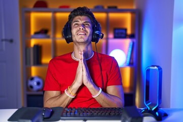 Young hispanic man playing video games begging and praying with hands together with hope expression on face very emotional and worried. begging.