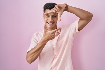 Young hispanic man standing over pink background smiling making frame with hands and fingers with happy face. creativity and photography concept.