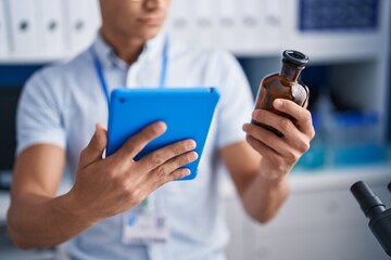 Young hispanic man scientist using touchpad holding bottle at laboratory