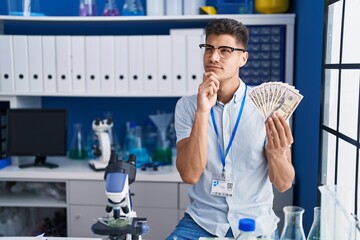 Young hispanic man working at scientist laboratory holding dollars serious face thinking about question with hand on chin, thoughtful about confusing idea