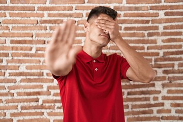 Young hispanic man standing over bricks wall covering eyes with hands and doing stop gesture with sad and fear expression. embarrassed and negative concept.